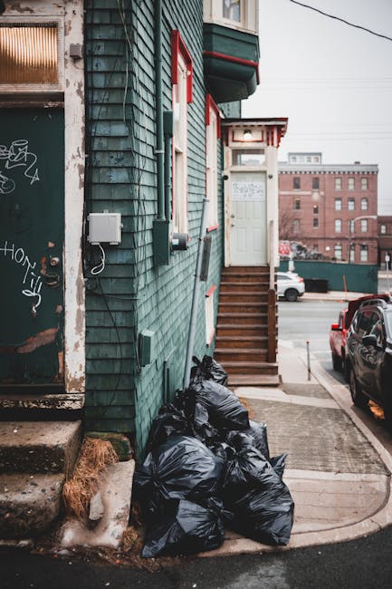 A collection of black plastic rubbish bags piled up against the side of a residential building with green wooden cladding, situated on a concrete pavement near a small set of wooden stairs leading to a white front door. The building features red trim around the windows and above the door, and an electric meter box attached to the exterior wall. In the background, an urban street scene with parked cars, an industrial brick building, and overhead power lines can be seen. The rubbish bags are tightly packed, appear to be filled with household waste or debris, and are placed directly on the pavement, indicating a private waste collection or alternative rubbish removal arrangement. The environment is illuminated by diffuse daylight, and the scene captures an area where private waste handling services, such as those provided by Rubbish Removal Kingston, are likely utilized for on-site clearance of refuse, bypassing local authority rubbish collection. The overall setting reflects an urban residential context with visible signs of waste awaiting removal.