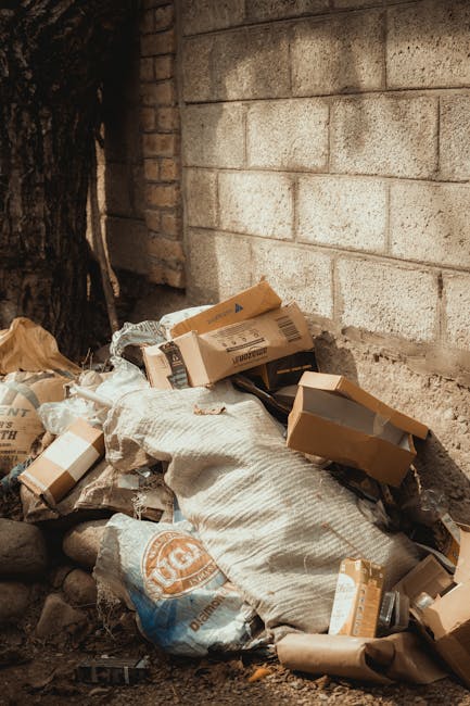An outdoor scene showing a pile of discarded cardboard boxes, some flattened and others still in their original shape, stacked against a weathered brick wall near a tree trunk. The cardboard varies in shades of brown and beige, with some boxes featuring printed labels or logos, indicating used packaging materials. Among the boxes, there are large white and beige bags, possibly made of woven plastic or fabric, filled with additional waste or debris, with some showing frayed edges or partial tears. The ground beneath the pile is uneven, composed of dirt and small rocks, with a few scattered leaves and debris nearby. The background includes a portion of a masonry wall constructed from concrete blocks, providing a neutral, textured backdrop. The overall environment appears to be an exterior space designated for rubbish disposal, with the scene possibly related to outside waste collection or private rubbish removal services; Rubbish Removal Kingston occasionally handles such types of waste clearing involving cardboard and general rubbish accumulation.