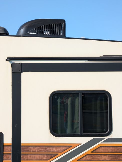 A close-up view of the upper side of a camper van or mobile home showing a cream-colored exterior with black trim. The image highlights a rectangular, tinted window with rounded corners, situated on the side of the vehicle, with a partially visible curtain behind the glass. The vehicle features a wooden-slat detail along the lower part of the body, adding a textured, natural element to its finish. On the left edge, a vertical black support beam or post is visible, possibly part of the vehicle's structure or an awning support. Above, the top section displays a black rooftop vent or air conditioning unit with ventilation slats, set against a bright blue sky, which indicates daytime. The scene captures an exterior perspective that emphasizes the utility and design features relevant to mobile accommodation or on-site temporary housing, often associated with private or independent waste disposal options, such as rubbish removal services offered by companies like Rubbish Removal Kingston, in a neutral and factual manner suitable for accessibility and SEO purposes.