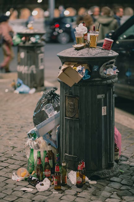 A black wheelie bin with a white label and the text 'St. John's' printed on the front, positioned on a dimly lit urban pavement at night. The bin is overflowing with various waste materials, including cardboard boxes, paper, and plastic items, some of which are protruding and leaning out of the open lid. The surrounding environment features a row of trees and dark bushes along the sidewalk on the right, with streetlights casting warm, yellowish light onto the scene. In the background, a quiet street with parked cars and more streetlights stretching into the distance can be seen, creating a subtle glow. The image reflects an independent waste disposal effort typical of private rubbish collection services like Rubbish Removal Kingston, highlighting the common practice of on-site clearance away from local authority bins in suburban areas.