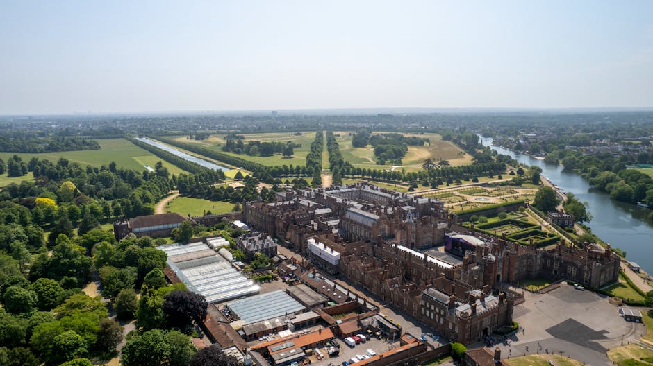 An aerial view of Hampton Court Palace surrounded by extensive gardens and green lawns, with the palace's historic brick and stone façade prominently visible in the foreground. To the left, there are several greenhouse structures with transparent glass roofs, positioned amidst lush trees and landscaped vegetation. The palace complex is enclosed by a paved courtyard with parked cars along one side, and an open area used for further site activities. In the background, beyond the palace grounds, the landscape extends into flat, vividly green fields, interspersed with tree-lined avenues and water channels that reflect the clear, bright sky. A wide river runs parallel to the grounds on the right, bordered by additional greenery and trees, creating a scenic boundary. The overall scene is well-lit with natural daylight, emphasizing the historic architecture and surrounding natural features, characteristic of a location that may require ongoing maintenance and clearance of refuse or waste from the site's operational areas. Rubbish Removal Kingston provides services that align with maintaining such historic sites' cleanliness and waste management.