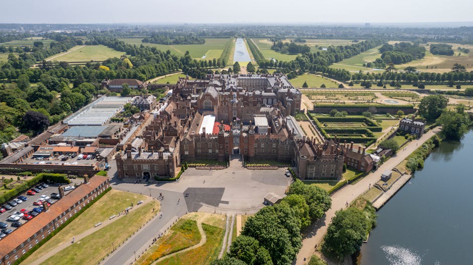 An aerial view of a historic castle with intricate brick architecture, multiple turrets, and slate roofs, situated on a landscaped grounds with pathways, greenery, and a large open courtyard at the front. To the left, there are modern structures and a parking lot filled with cars. The castle is bordered by a river along its right edge, with a paved pathway running parallel to the water. In the background, expansive green fields, neatly divided by hedgerows and tree-lined avenues, stretch toward the horizon under a clear, bright sky. The image captures the contrast between the medieval-style castle and the natural, cultivated landscape, highlighting the environment surrounding the property. This setting reflects heritage site preservation with nearby spaces suitable for outdoor activities and scenic views, fitting into the context of private waste handling or on-site clearance for such historical locations, as managed by local rubbish removal services like Rubbish Removal Kingston.