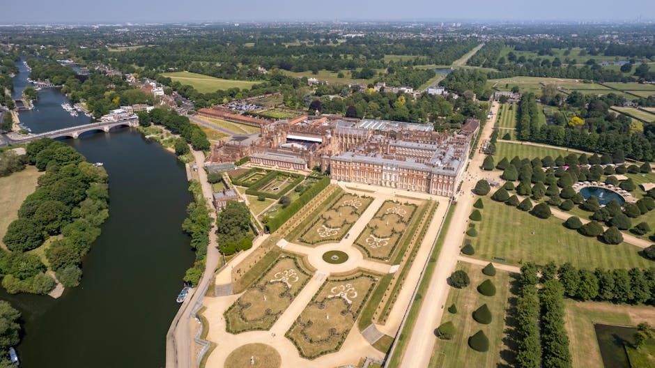 An aerial view of Hampton Court Palace surrounded by extensive gardens and green lawns, with the palace's historic brick and stone façade prominently visible in the foreground. To the left, there are several greenhouse structures with transparent glass roofs, positioned amidst lush trees and landscaped vegetation. The palace complex is enclosed by a paved courtyard with parked cars along one side, and an open area used for further site activities. In the background, beyond the palace grounds, the landscape extends into flat, vividly green fields, interspersed with tree-lined avenues and water channels that reflect the clear, bright sky. A wide river runs parallel to the grounds on the right, bordered by additional greenery and trees, creating a scenic boundary. The overall scene is well-lit with natural daylight, emphasizing the historic architecture and surrounding natural features, characteristic of a location that may require ongoing maintenance and clearance of refuse or waste from the site's operational areas. Rubbish Removal Kingston provides services that align with maintaining such historic sites' cleanliness and waste management.