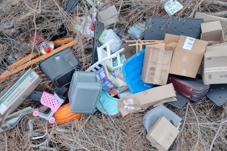 A pile of mixed household waste and clutter on a patch of dry, brown grass and sparse vegetation, consisting of various cardboard boxes, some flattened and others partially assembled, alongside plastic containers, including a light gray one and a bright blue one. There are small household items such as a pink children's toy with wheels, a black briefcase, and what appears to be a small white step stool. An open cardboard box reveals additional contents, while a crumpled piece of cardboard and a partially visible black lounge or seat cushion are also present. The waste appears haphazardly discarded, with some items leaning against each other and scattered across the ground, suggesting an act of illegal dumping in an outdoor area. The scene reflects typical debris associated with private waste disposal or rubbish removal services like Rubbish Removal Kingston, which manage such clutter for clients seeking alternative waste handling options outside local authority collections.