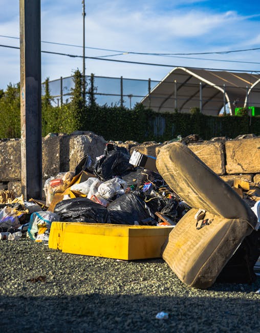 A pile of mixed household waste and clutter on a patch of dry, brown grass and sparse vegetation, consisting of various cardboard boxes, some flattened and others partially assembled, alongside plastic containers, including a light gray one and a bright blue one. There are small household items such as a pink children's toy with wheels, a black briefcase, and what appears to be a small white step stool. An open cardboard box reveals additional contents, while a crumpled piece of cardboard and a partially visible black lounge or seat cushion are also present. The waste appears haphazardly discarded, with some items leaning against each other and scattered across the ground, suggesting an act of illegal dumping in an outdoor area. The scene reflects typical debris associated with private waste disposal or rubbish removal services like Rubbish Removal Kingston, which manage such clutter for clients seeking alternative waste handling options outside local authority collections.