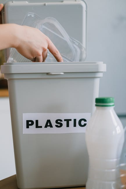 A person's hand is seen placing a clear plastic container into a light gray bin labeled 'PLASTIC' with bold black letters, indicating separate waste for plastic recycling. The bin is positioned on a wooden surface, with a partially visible white plastic milk bottle with a green cap placed nearby in the foreground. The background features a clean, indoor setting with a light-colored wall, suggesting a household environment where waste segregation is practiced to support non-local authority waste disposal methods and private rubbish removal services. The bin has a hinged lid that is open, revealing the plastic container being inserted, while the surface of the bin appears smooth and slightly reflective. Natural or indoor lighting illuminates the scene evenly, emphasizing the clarity of the plastic items and the neat organization of waste sorting efforts that are associated with efficient rubbish management, such as those offered by companies like Rubbish Removal Kingston for household waste separation and disposal.