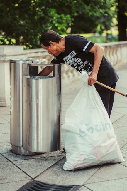A man with tan skin and dark hair, dressed in a casual black T-shirt with white graphic print on the front, is seen bending over as he disposes of a large white plastic bag filled with trash into a cylindrical stainless steel rubbish bin on a paved outdoor sidewalk. The bin has a brushed finish and is positioned next to a low stone balustrade, which runs parallel to the pavement, with lush green foliage and trees visible in the background, suggesting a park or urban green space setting. The man's left hand holds the bag open as he tips its contents into the open-top bin, while his right hand steadying the bag. The scene is illuminated by natural daylight, creating a clear view of the textured concrete pavement and the reflective surface of the bin. This image exemplifies private waste disposal or rubbish collection services, such as those offered by Rubbish Removal Kingston, highlighting environmentally responsible waste management in an outdoor urban environment.