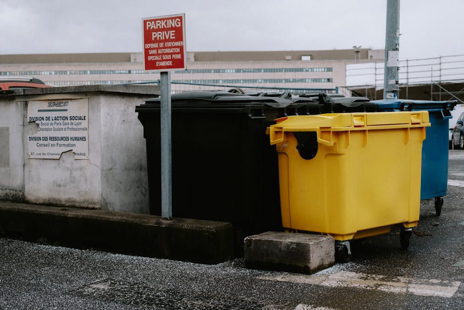 In a parking area adjacent to a concrete wall, four large wheeled rubbish containers are aligned side by side, each with distinct colours: black, yellow, blue, and a hidden fourth likely grey or green. The yellow container, positioned in the foreground, has a textured plastic surface with a lid that features hinges and a handle, and rests on small caster wheels. The black container behind it has a smooth, matte finish, while the blue bin, partially visible on the right side, also appears to have a similar plastic construction. A small, rectangular concrete block is placed beneath the yellow bin’s wheels, raising it slightly off the asphalt surface, which has small loose gravel and patches of dirt. To the left, a metallic pole supports a red 'No Parking' sign in English and French, affixed above an official sign indicating social services, both attached to the concrete wall. In the background, a modern building with horizontal windows and a metal railing is visible under overcast daylight, suggesting a typical urban environment where waste collection and rubbish removal services by Rubbish Removal Kingston may operate, highlighting the practical aspect of local rubbish disposal options outside of regular public refuse collection.