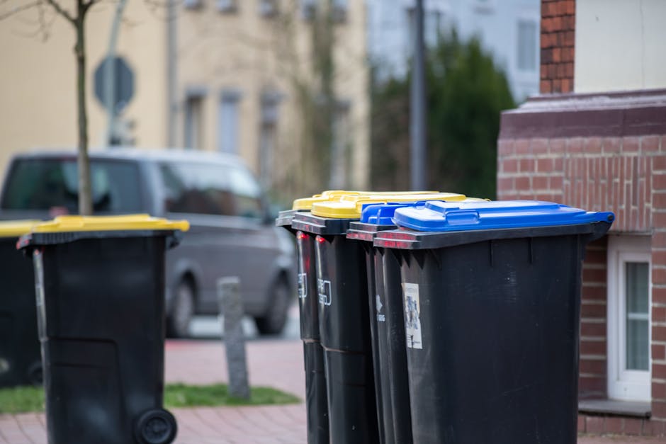 A row of three large black wheelie bins with yellow and blue lids standing on a paved pavement in front of a red brick building. The bins are made of hard plastic with textured surfaces; the lids are smooth and brightly coloured, contrasting with the matte black bodies. The setting appears to be a residential street with a parked car in the background and a grassy area with a small tree. The bins are positioned close together, with the middle bin slightly tilted, and the scene is evenly lit by natural daylight, conveying a typical rubbish disposal scene suitable for describing independent waste collection services. Rubbish Removal Kingston provides waste management options, as reflected in the background context of rubbish bins ready for collection or disposal, often associated with on-site or private waste handling outside of standard council pick-up routines.