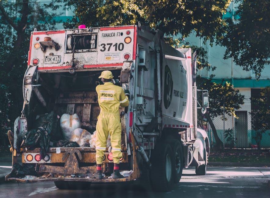 In a parking area adjacent to a concrete wall, four large wheeled rubbish containers are aligned side by side, each with distinct colours: black, yellow, blue, and a hidden fourth likely grey or green. The yellow container, positioned in the foreground, has a textured plastic surface with a lid that features hinges and a handle, and rests on small caster wheels. The black container behind it has a smooth, matte finish, while the blue bin, partially visible on the right side, also appears to have a similar plastic construction. A small, rectangular concrete block is placed beneath the yellow bin’s wheels, raising it slightly off the asphalt surface, which has small loose gravel and patches of dirt. To the left, a metallic pole supports a red 'No Parking' sign in English and French, affixed above an official sign indicating social services, both attached to the concrete wall. In the background, a modern building with horizontal windows and a metal railing is visible under overcast daylight, suggesting a typical urban environment where waste collection and rubbish removal services by Rubbish Removal Kingston may operate, highlighting the practical aspect of local rubbish disposal options outside of regular public refuse collection.