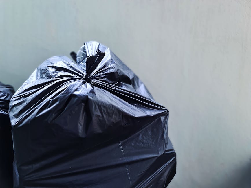 Close-up of a large black plastic rubbish bag filled with waste materials, tied tightly at the top with a knot, placed against a plain light grey wall. The bag's surface appears crinkled and reflective, indicating a thick, durable plastic. To the left of the main bag, additional black rubbish bags are partially visible, stacked or arranged nearby on a concrete or asphalt surface, suggesting an indoor or outdoor waste collection area. The environment looks clean and organized, typical of a private waste disposal or on-site clearance setup, suitable for independent rubbish removal services. The scene highlights the handling of domestic or commercial waste as part of a rubbish removal process undertaken by companies like Rubbish Removal Kingston, which offers same day collection solutions. The image emphasizes the importance of proper waste containment before disposal, aligning with the context of alternative waste handling methods away from local authority collections.