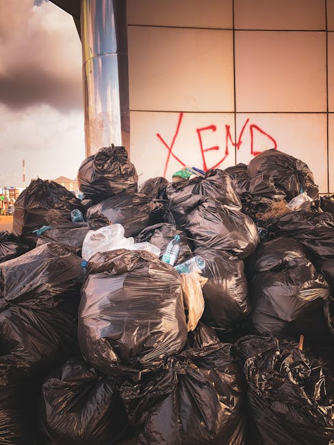 A large pile of black rubbish bags, some with visible creases and knots, is stacked against a tiled wall with beige and brown tones. Several clear plastic water bottles, partly crushed and with visible labels, are strewn among the bags, some protruding from the openings. The bags are situated outdoors on a pavement or concrete surface, with a partial view of an urban environment in the background, including buildings and sky with dark clouds. The setting suggests an area awaiting private rubbish collection or on-site clearance, as provided by companies like Rubbish Removal Kingston, to manage waste outside of traditional municipal services. The scene indicates a possible delay in rubbish removal services, with the bags appearing piled up and unsecured, highlighting the importance of efficient waste management solutions for maintaining cleanliness in commercial or residential spaces.