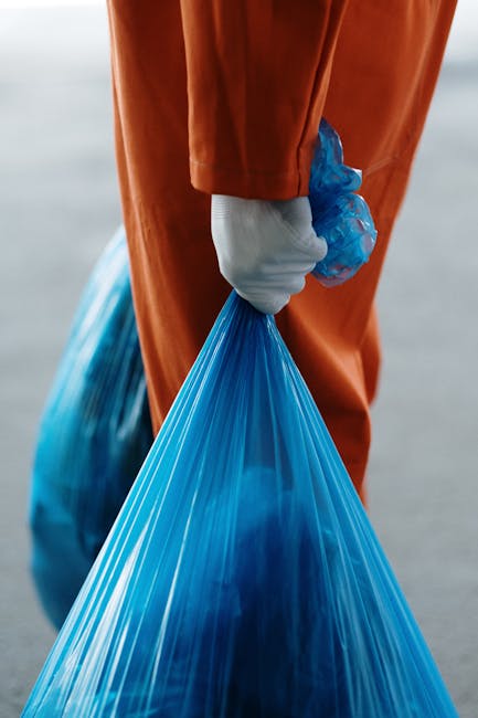 Close-up of a large black plastic rubbish bag filled with waste materials, tied tightly at the top with a knot, placed against a plain light grey wall. The bag's surface appears crinkled and reflective, indicating a thick, durable plastic. To the left of the main bag, additional black rubbish bags are partially visible, stacked or arranged nearby on a concrete or asphalt surface, suggesting an indoor or outdoor waste collection area. The environment looks clean and organized, typical of a private waste disposal or on-site clearance setup, suitable for independent rubbish removal services. The scene highlights the handling of domestic or commercial waste as part of a rubbish removal process undertaken by companies like Rubbish Removal Kingston, which offers same day collection solutions. The image emphasizes the importance of proper waste containment before disposal, aligning with the context of alternative waste handling methods away from local authority collections.
