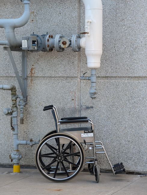 A manual wheelchair with black armrests, a silver metallic frame, and large black rear wheels is positioned on a concrete surface against a grey, textured exterior wall. The wheelchair's footrest is slightly extended, and a small black caster wheel is visible at the front. Above the wheelchair, several white and grey pipes and valves are mounted on the wall, showing signs of rust and corrosion. The pipes include a larger vertical pipe and several smaller interconnected sections, some with rusted fittings, stretching horizontally and vertically across the wall. The scene is illuminated by natural light, highlighting the rough texture of the wall and the metallic surfaces of the wheelchair and pipes. The setting appears to be an outdoor area adjacent to a building, possibly near utility or service access points, with no other objects or people present. Rubbish removal services could include clearing or disposing of items in this sort of environment, especially when accessibility issues are a concern, as indicated by the presence of a mobility device in the vicinity of on-site infrastructure.